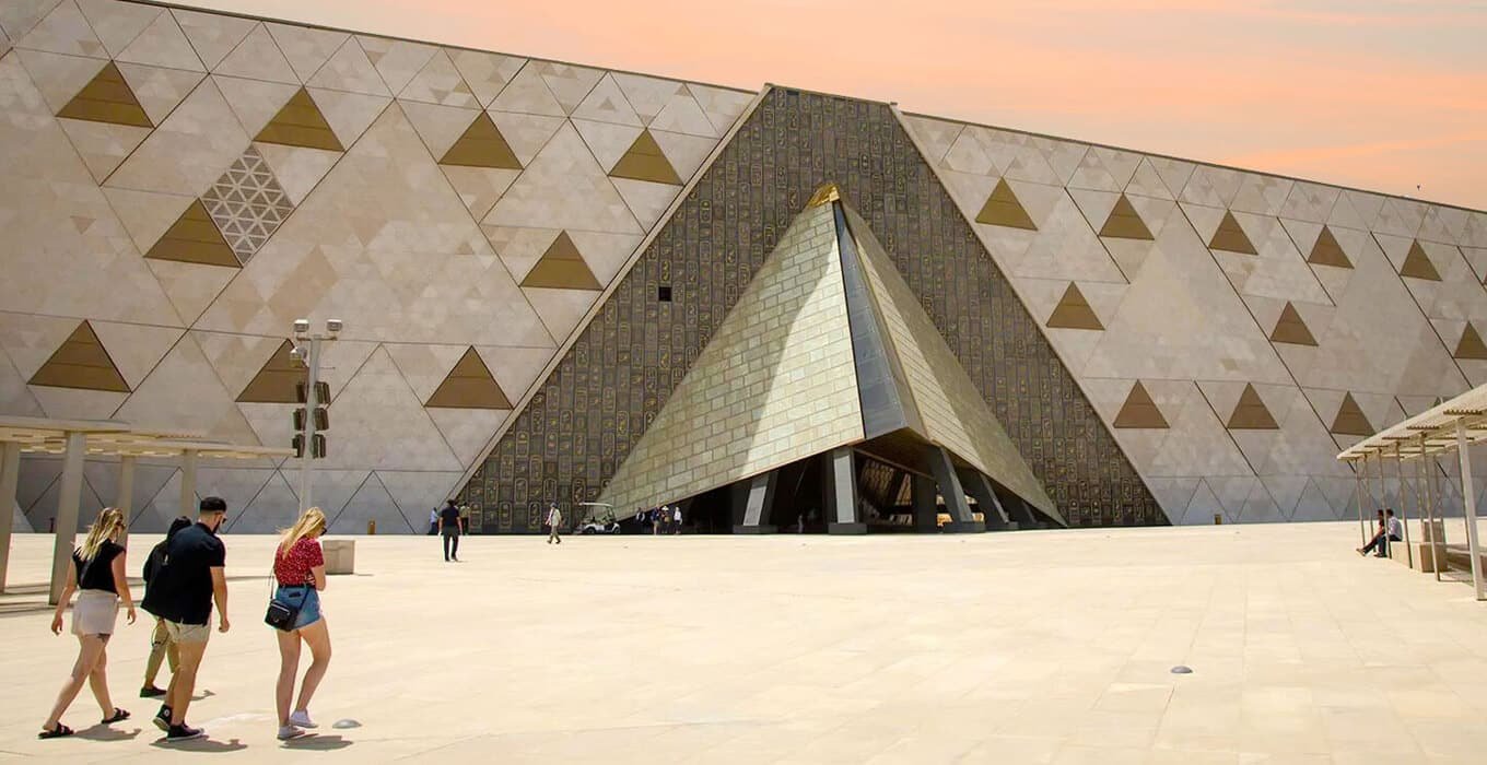 Modern Grand Egyptian Museum building entrance with large triangular stone wall and visitors walking across plaza
